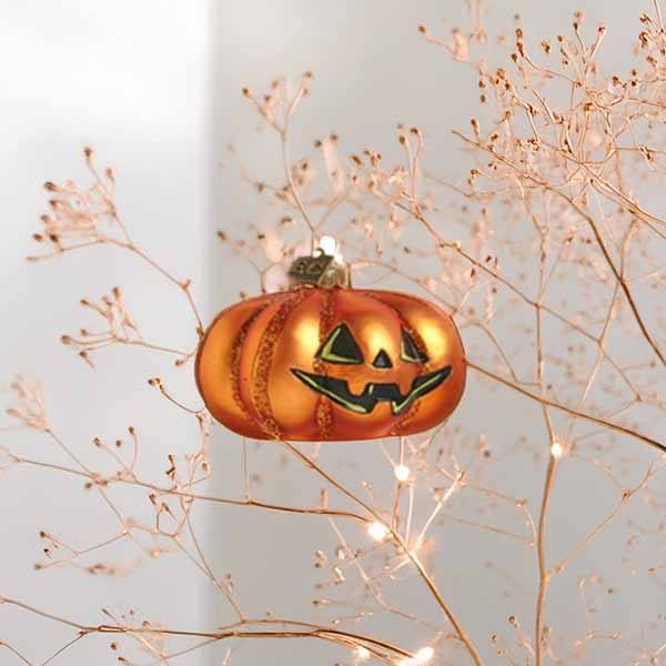 The Ornament - Pumpkin Jack-o-Lantern from Available for Pick-up & Delivery features a glass pumpkin with a black, smiling face, hanging from lit branches to create a festive Halloween atmosphere.