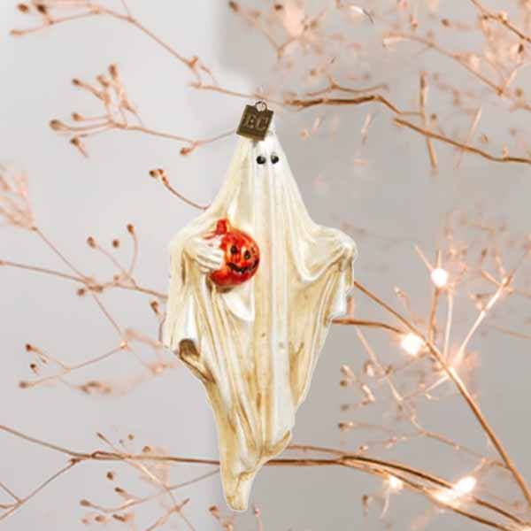 The Ornament - Friendly Ghost by Available for Pick-up & Delivery showcases a friendly ghost with black eyes, wrapped in cream fabric, clutching a red jack-o-lantern. It elegantly hangs from lit branches, casting a whimsical glow.