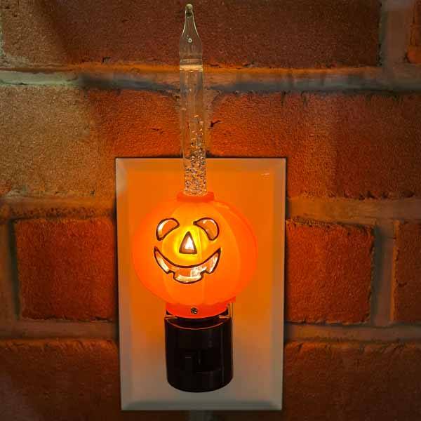 The Pumpkin Bubble Light by Available for Pick-Up & Delivery plugs into a wall socket, emitting a warm orange glow with its candle-like top. Set against a brick wall, it offers a vintage touch for a cozy Halloween-themed ambiance.
