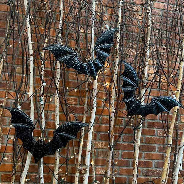Three artificial Bat - Decor from Available for Pick-Up & Delivery hang from bare tree branches against a brick wall, adorned with small white lights to create an elegant and spooky Halloween atmosphere.