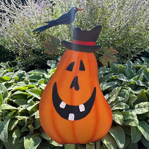 The Jack-o-Lantern with Hat & Crow Pumpkin Stake – 28” from Available for Pick-Up & Delivery features a cheerful pumpkin with a black hat and crow, set off beautifully by lush green leaves and tall plants, making it an inviting Halloween garden decor piece.