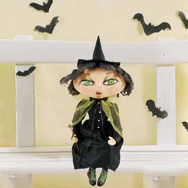 A 23 Xanzabelle Witch doll by Joe Spencer, part of the Available for Pick-Up & Delivery brand, sits on a white shelf with a black hat and green cape. Black paper bats adorn the cream wall, enhancing the spooky Halloween decor.