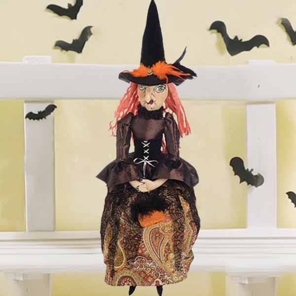 The 36 Elfreda Witch from Available for Pick-up & Delivery, with red hair and a tall black hat, sits on a white shelf in her paisley-patterned dress. Bats cast shadows on the pale yellow wall behind, enhancing this enchanting handcrafted figures spooky allure.