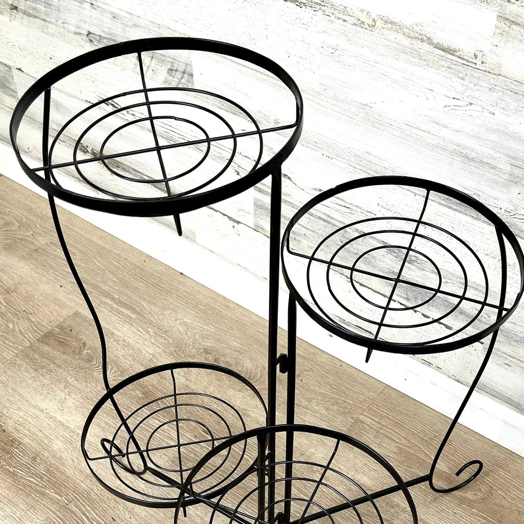 The Plant Stand – Black Metal Folding – 4 Tier by Pickup & Local Delivery Only features a sleek, minimalist geometric design, perfect for modern interiors, and is shown on a light wood floor against a white textured wall.