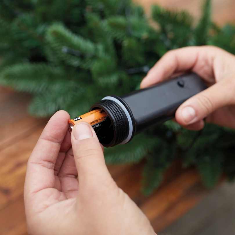 A person inserts an AA battery into a black compartment to power Durawise Battery Operated LED Twinkle String Lights Warm White by Available for Pickup & Delivery, arranging them in green pine branches for festive holiday décor.