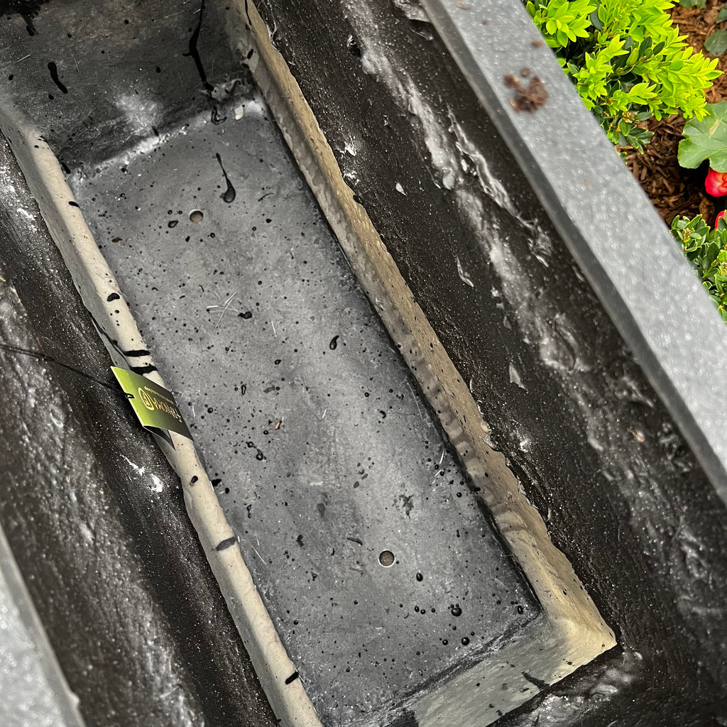 A close-up of the empty Blackburn Divider Planter (32x12, black) by Pick-Up & Local Delivery Only shows its drainage holes, a plant tag on the edge, and green foliage with flowers in the background.
