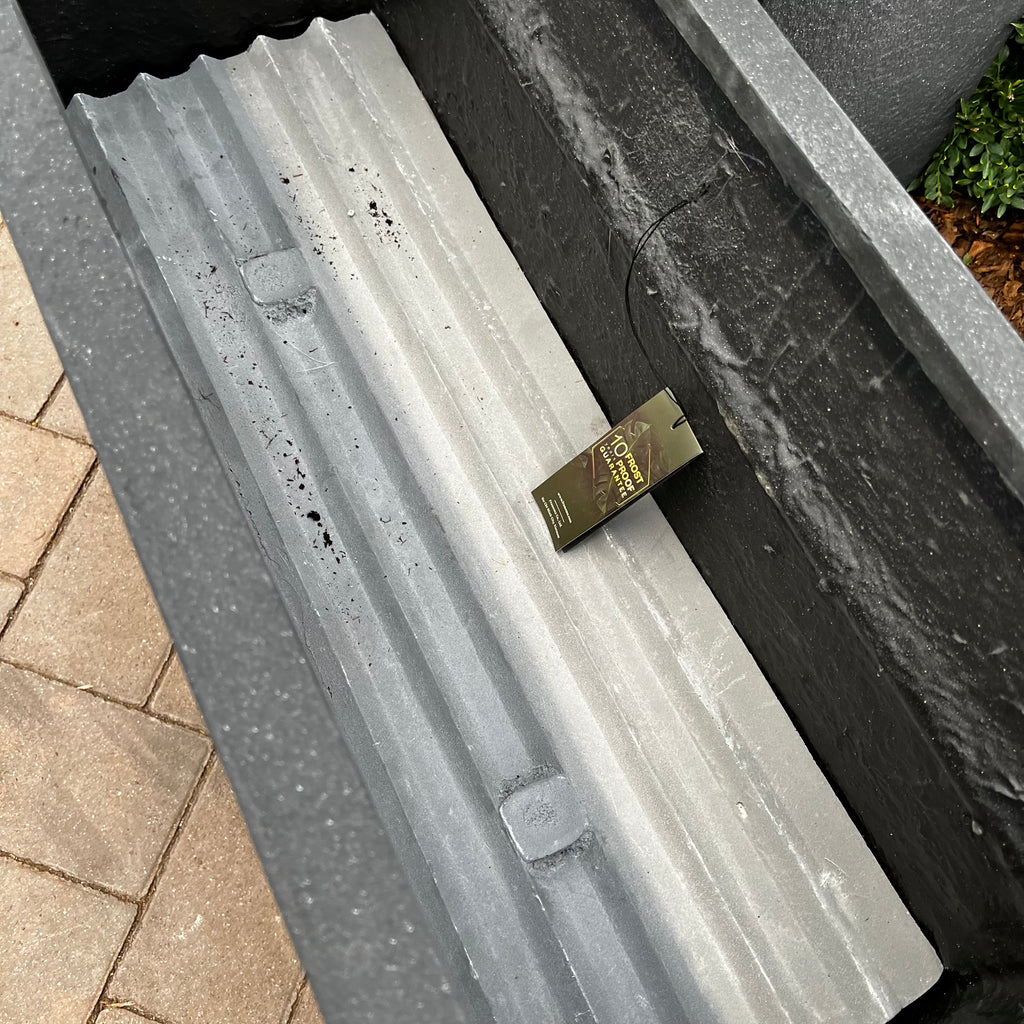 The Blackburn Divider Planter (32x12) by Pick-Up & Local Delivery Only in black features a gray corrugated liner and plant tag, making it perfect for garden or patio divider use on paved surfaces.