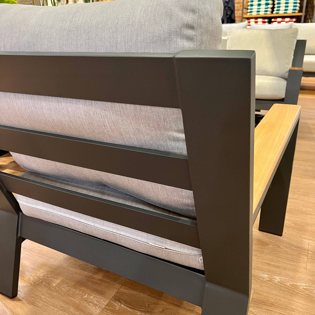 Close-up of a Bozania 3-Piece Outdoor Patio Seating Set armchair by Delivery Only, with matte black metal frame, light gray cushions, and light wood armrest. A similar chair appears in the background—ideal for stylish outdoor lounging.