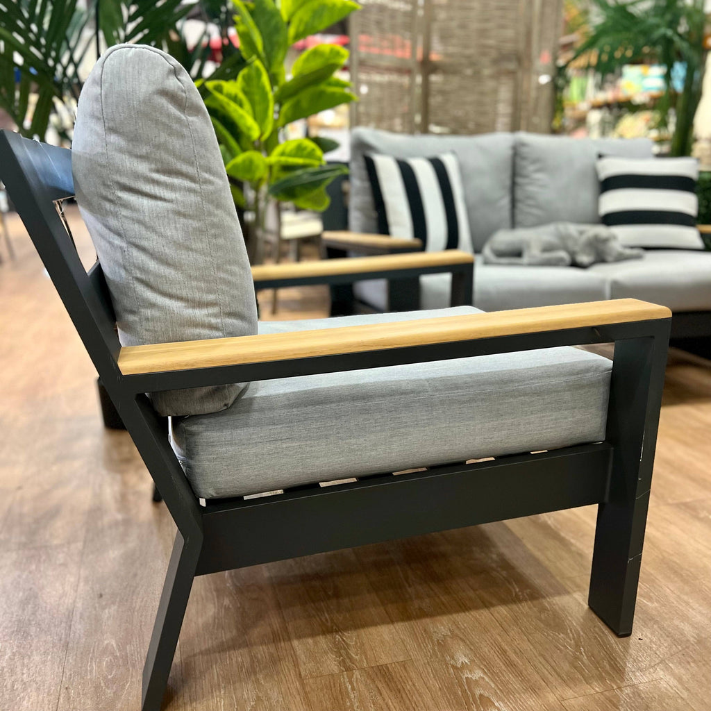 A modern outdoor lounge chair with light gray cushions and wooden armrests sits on a wood floor. In the background, the Delivery Only Bozania 3-Piece Outdoor Patio Seating Set with striped pillows complements green plants.