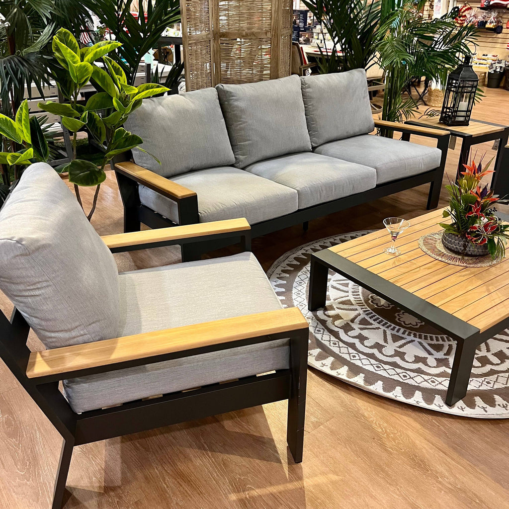 Create a contemporary indoor look with the Delivery Only Bozania 3-Piece Outdoor Patio Seating Set—gray cushioned sofa, armchair, wood coffee table, and decor like plants and a floral centerpiece. A martini glass sits on the table.