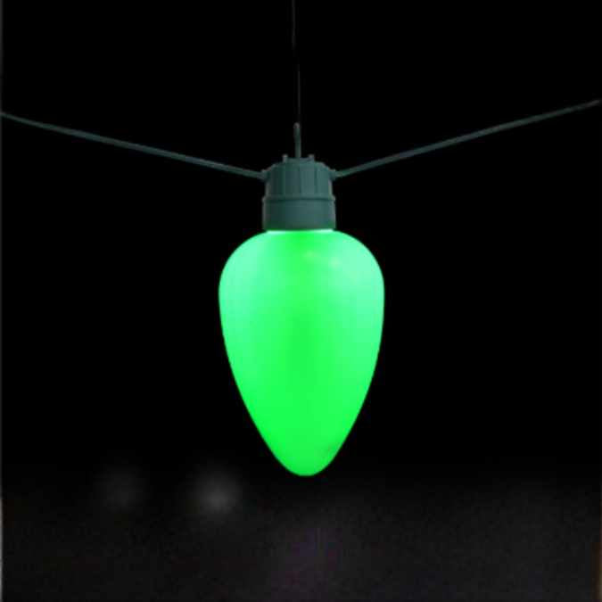 The Jumbo LED Lit Bulb - Green by Available for Pick-Up & Delivery, shaped like a retro C9 bulb, hangs against a black background with two wires extending diagonally from the top.