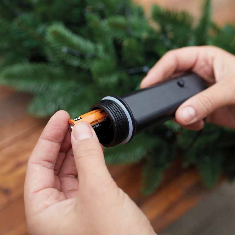A person inserts an AA battery into a black compartment to power the Durawise Battery Operated LED Twinkle String Lights – Warm White, by Available for Pick-Up & Delivery, with green artificial pine branches in the background.