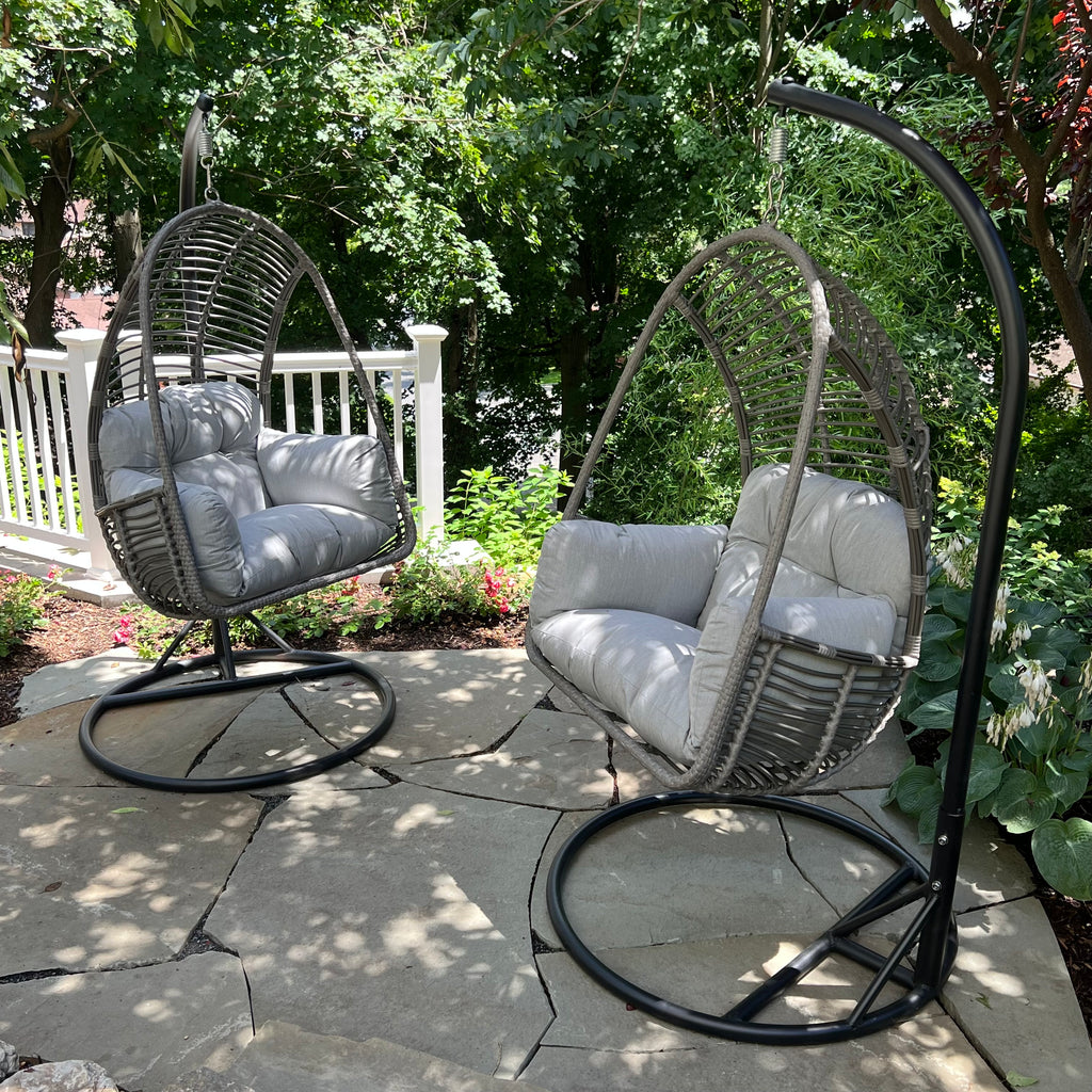 Two Pick-Up & Local Delivery Only Hanging Basket Chairs with grey cushions and durable metal frames are on a stone patio surrounded by greenery, flowers, and a white railing in the background.
