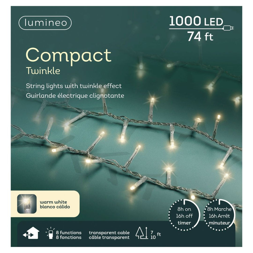 A close-up of LED 1000 ct. Compact String Lights Twinkle Warm White by Available for Pick-Up & Delivery on a greenish background. The 74 ft box highlights 8 functions, timer, transparent cable—perfect for outdoor holiday decor. English and French text shown.