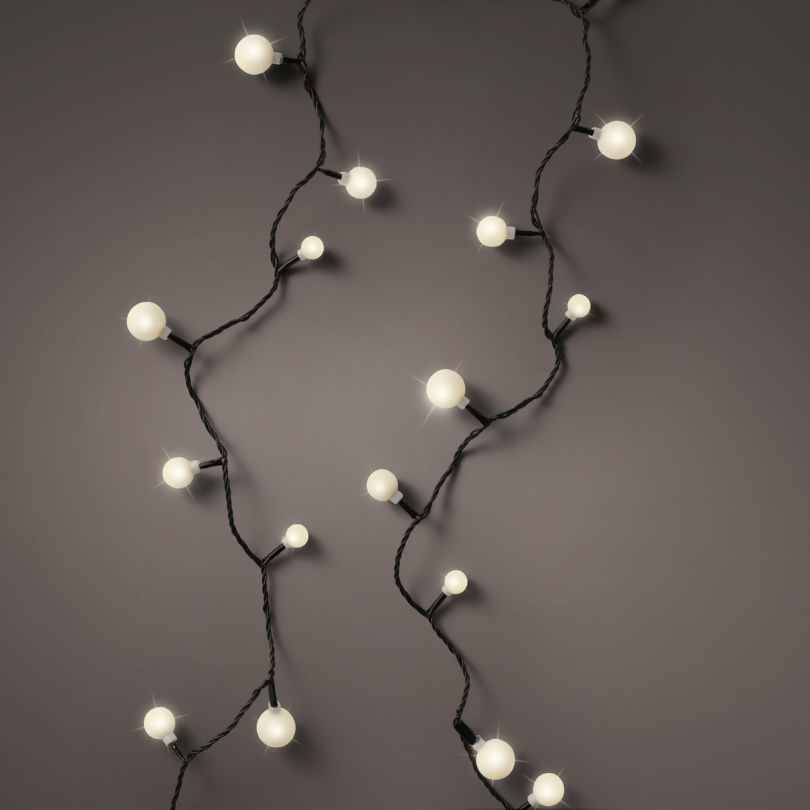 The Cherry LED 120 ct. String Lights Warm White by Available for Pickup & Delivery hang against a dark gray wall, their round bulbs on black cords casting a soft, cozy glow ideal for holiday décor.