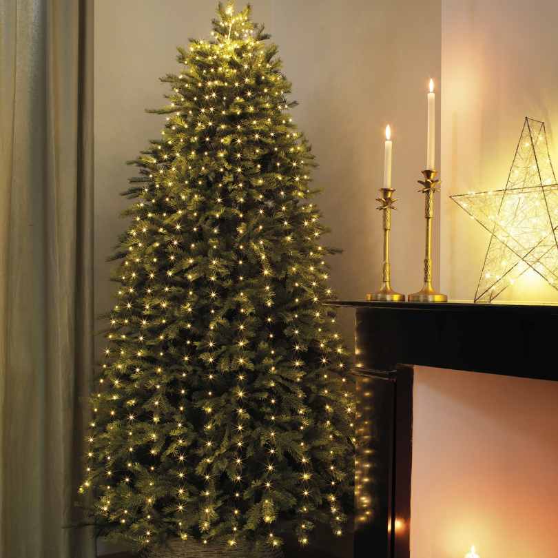 A Micro LED 672 ct. Tree Bunch Warm White by Available for Pick-Up & Delivery stands near the fireplace, complemented by gold candle holders and a glowing star on the mantel for a cozy holiday atmosphere.