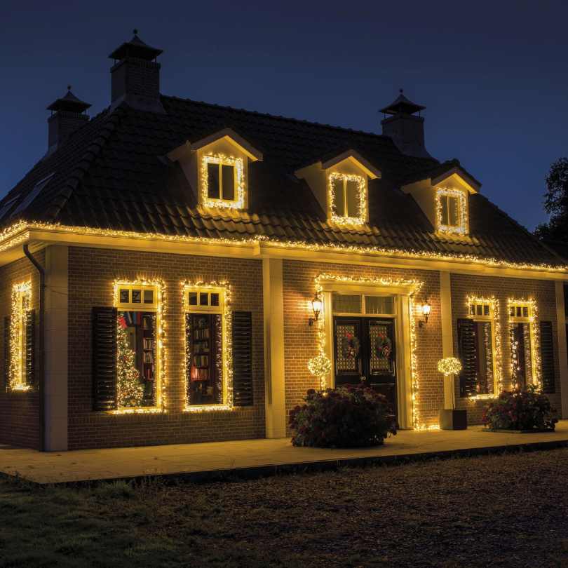 A house adorned with the Compact Twinkle LED 500 ct. String Light Set Warm White from Available for Pickup & Delivery glows softly at night, outlining the roof, windows, and doors against the dark sky.