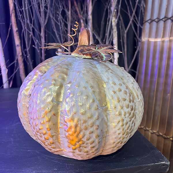 The Pumpkin - Metal - Round by Available for Pick-Up & Delivery, featuring a textured metallic surface and gold-toned stem and leaf accents, embodies fall decor. It sits on a dark background with branches forming a rustic backdrop to this autumnal scene.