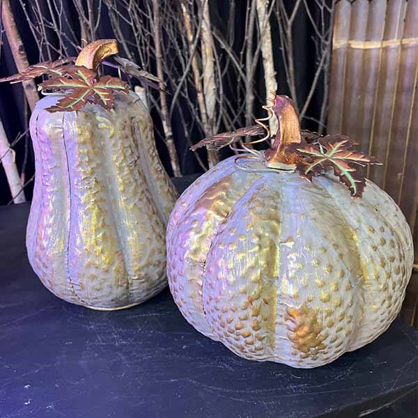 Two metal pumpkins with embossed textures and autumn leaves, set against a dark, rustic backdrop with branches. One is elongated with a gold stem, and the other is round. Ideal for fall decor. Brand: Available for Pick-Up & Delivery; Product: Pumpkin - Metal - Round.