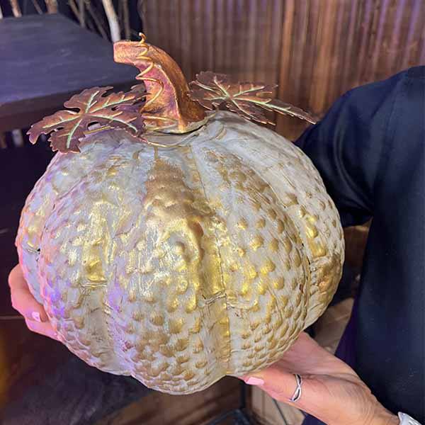 A person holds a decorative metal pumpkin from Available for Pick-Up & Delivery, perfect for fall decor. Its gold and silver textured surface features metallic leaves and a shiny gold stem, with a rustic wooden backdrop blurred in the background.