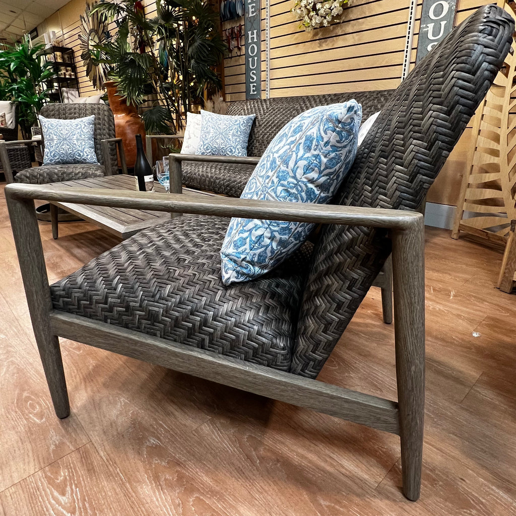 An inviting outdoor space includes the Provence 4 pc. Sofa, Lounge Chair, Swivel Chair, and Coffee Table Set by Delivery Only, featuring dark woven designs. Blue floral pillows add vibrancy, while potted plants and wooden wall decor enhance the setting of this all-weather wicker arrangement.