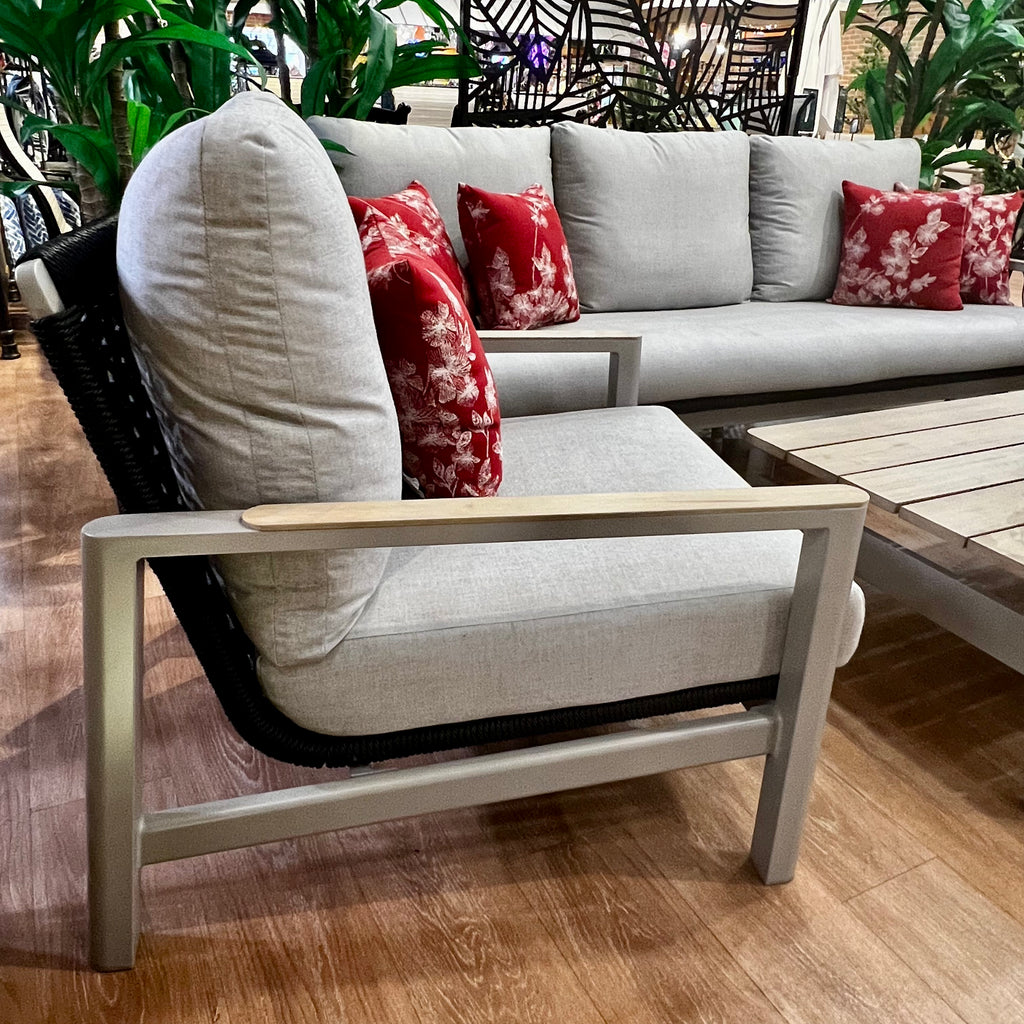 Enhance your modern indoor space with the Delivery Only Titan 4-Piece Seating Set. It includes a gray-cushioned outdoor sofa, lounge chair, swivel glider, and wooden coffee table adorned with red floral pillows amidst lush plants on a wooden floor for ultimate comfort.