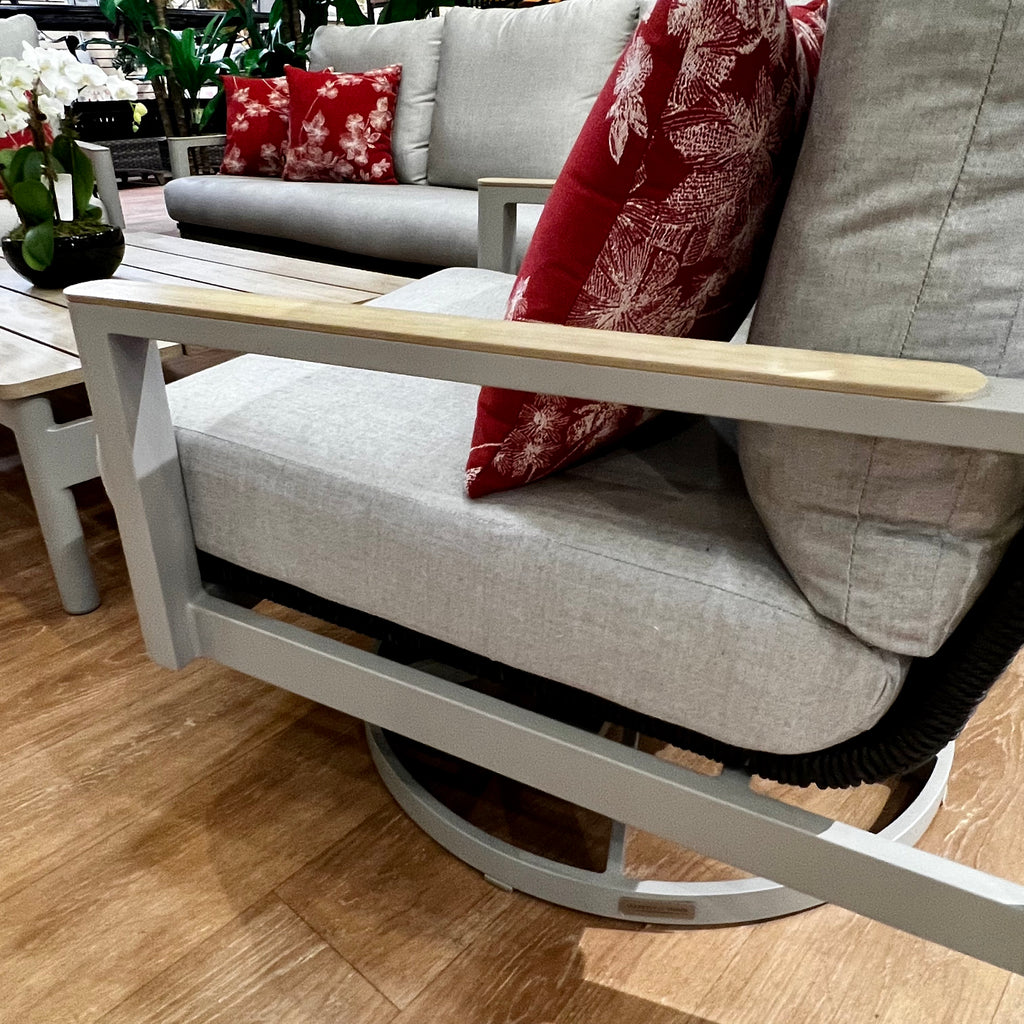 The Delivery Only Titan Outdoor Patio Set includes a sofa, lounge chair, swivel glider, and coffee table. Gray cushioned chairs feature wooden armrests with red pillows of a white leaf pattern, perfect for any outdoor space with wooden floors and lush plants.