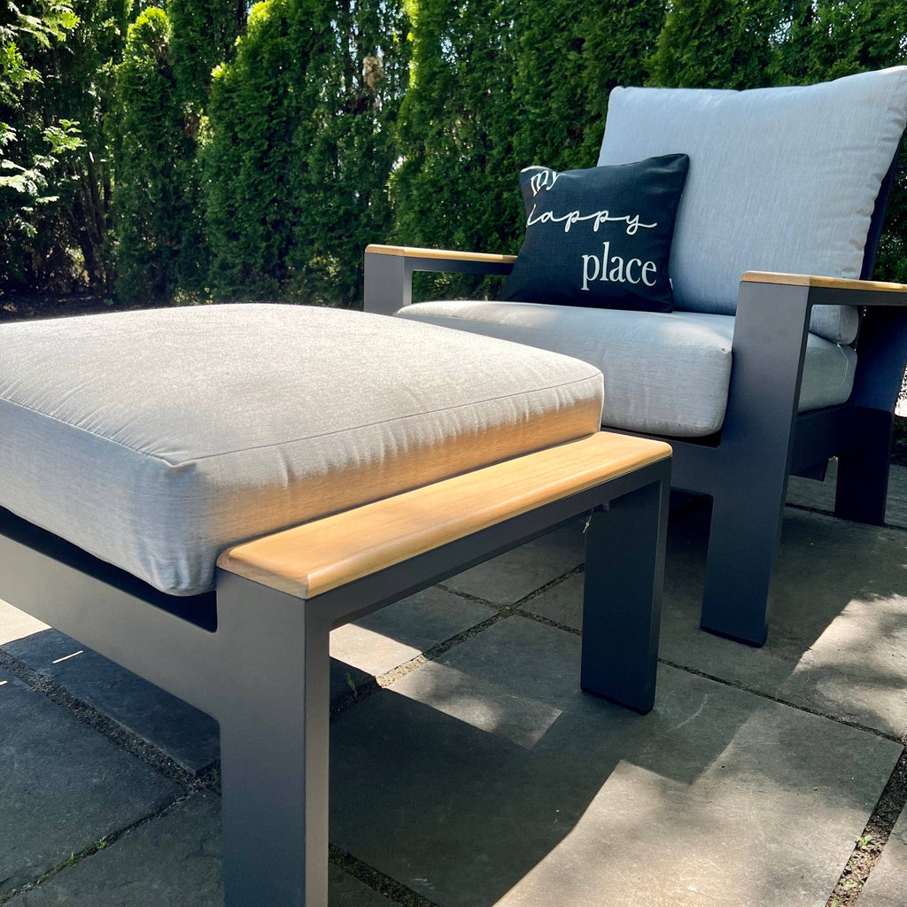 The Delivery Only Bozania Patio Lounge Chair Recliner features Sunbrella cushions on a stone patio, with My happy place on the black cushion. The chair, surrounded by greenery, includes adjustable reclining positions and is finished with black frames and wooden armrests.