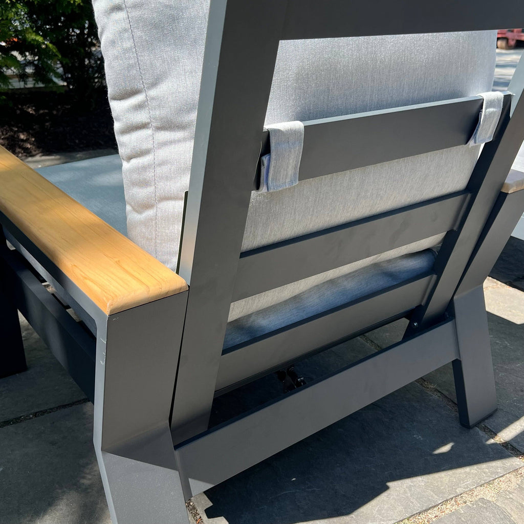 Rear view of the Bozania Patio Lounge Chair Recliner by Delivery Only, featuring gray Sunbrella cushions, wooden armrests, a sturdy modern metal frame on a stone patio; sunlight and nearby plants enhance its serene setting.