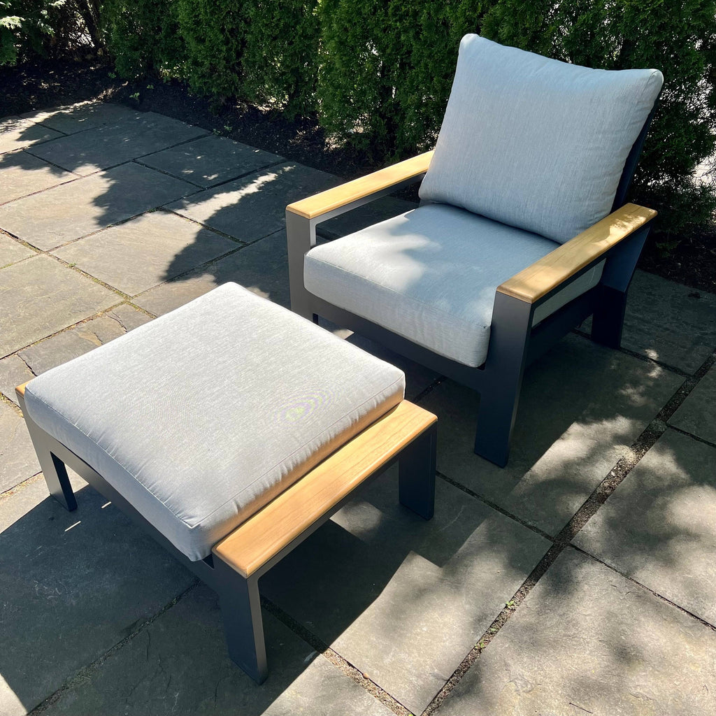A Bozania Patio Lounge Chair Recliner from Delivery Only features an adjustable recline and a matching ottoman on a stone floor. Both have light gray Sunbrella cushions and wooden armrests, with sunlight casting shadows and green bushes in the background.