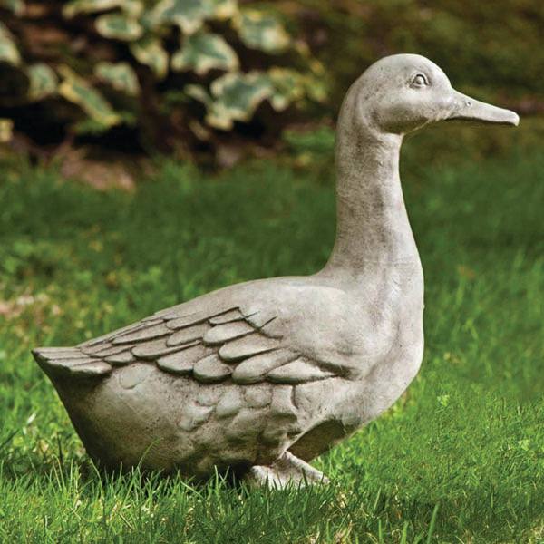 The All Delivery Options Statue – Cast Stone – Duck features detailed feathers and a raised head, creating a charming accent on green grass with softly blurred leafy plants in the background.