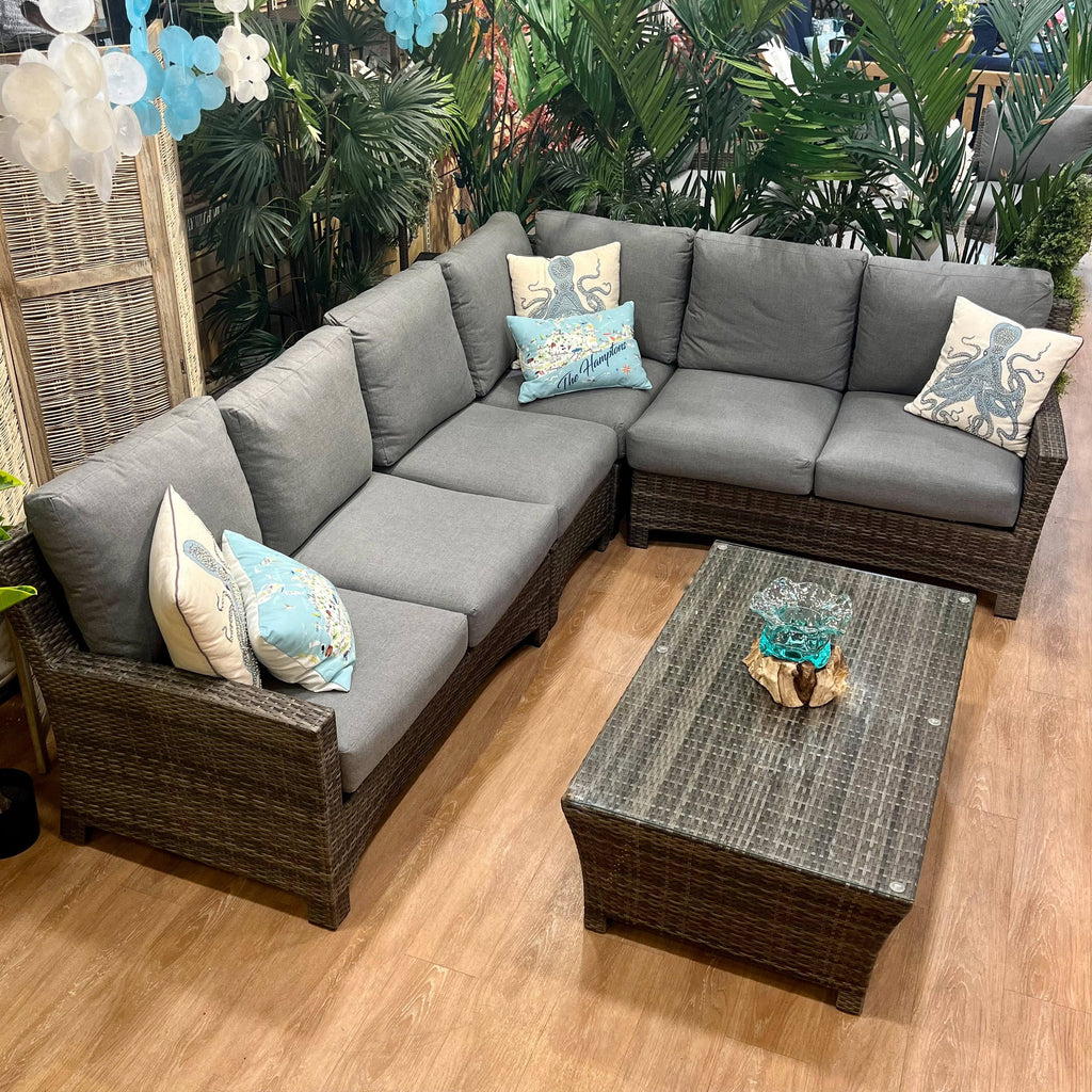 The Delivery Only Key Largo 4-Piece Outdoor Patio Sectional Seating Set features all-weather wicker, gray Sunbrella cushions, sea-themed pillows, and a matching glass-top coffee table on a wooden floor amid lush greenery.