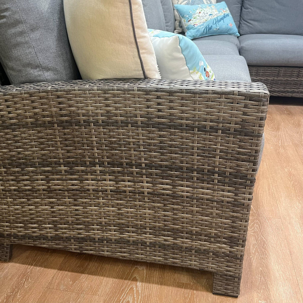 A close-up of Delivery Only’s Key Largo 4-Piece Outdoor Patio Sectional Seating Set shows its all-weather wicker arm with grey Sunbrella cushions and decorative pillows on a wooden floor, with more cushions and a light blue pillow in the background.