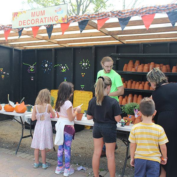 Children and an adult enjoy a Mum Decorating activity with crafting supplies and potted plants. Nearby, a sand art station contributes to the fun as a man in green assists them. Festive decorations hang above, all celebrating the day with Pick-Up Onlys Fall Festival Wristbands.