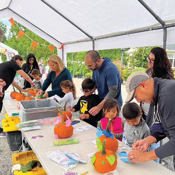 Children and adults decorate pumpkins with colorful paper under a tent at the Fall Festival Wristband booth from Pick-Up Only, where art supplies fill the table and small orange flags create a festive atmosphere.