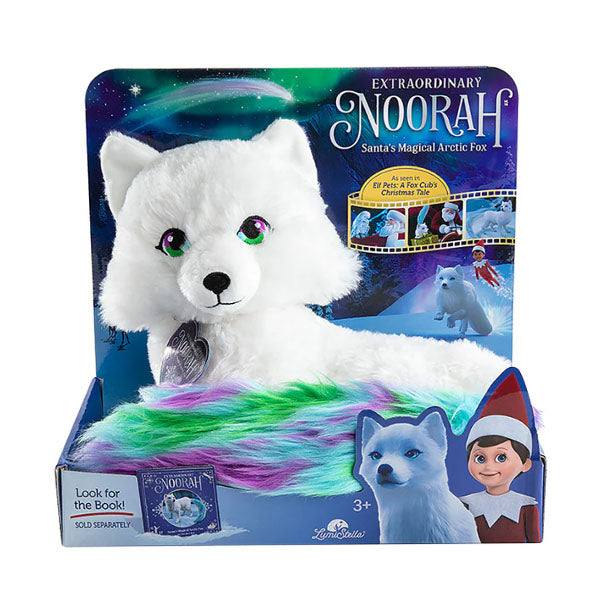 Noorah, an arctic fox plush toy in the display box. She has white fir and a green, blue and purple colored tail.