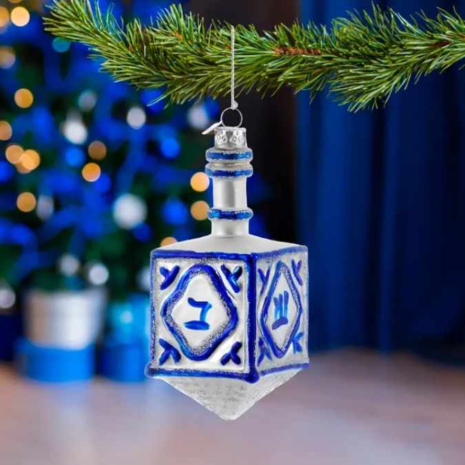 The Ornament - Dreidel by Available for Pick-Up & Delivery hangs from a pine branch, with blue and white holiday lights in the background—a beautiful interfaith decoration for your home.