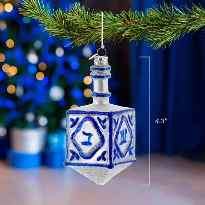 The Ornament - Dreidel by Available for Pick-Up & Delivery is a 4.3-inch silver and blue glass ornament hanging from a pine branch, with a blurred blue and white tree behind it—perfect as an interfaith holiday decoration.