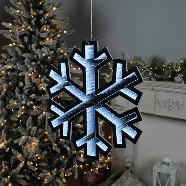 The Infinity Light – Snowflake – 23.5 glows indoors against a lit Christmas tree, capturing winters beauty. A mantel decorated with greenery enhances this festive holiday decor. Available for Pick-Up & Delivery.