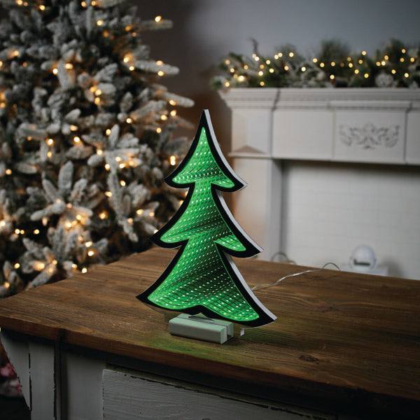 A 12” Infinity Light tree from Available for Pick-Up & Delivery glows on a wooden table. In the background, a larger tree sparkles with white lights, surrounded by holiday décor, while a mantel is adorned with greenery and features an enchanting infinity light display.
