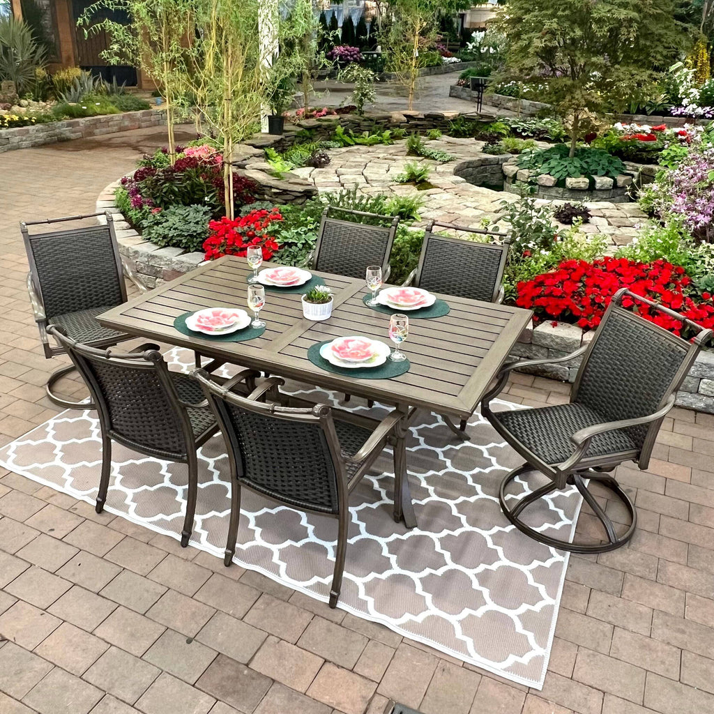 The Morena 7-piece Outdoor Patio Dining Set by Delivery Only features six chairs and a rectangular table for four, arranged on a stylish outdoor rug amid vibrant flowers, lush greenery, and a stone garden backdrop.