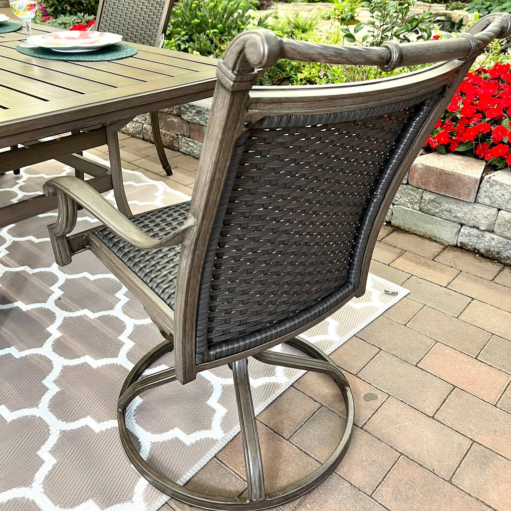 The Morena 7-piece Outdoor Patio Dining Set by Delivery only includes a wicker chair with a metal frame, placed on a patterned rug on a brick patio, surrounded by red flowers and greenery.