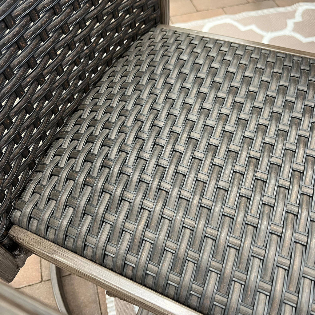 Close-up of the Morena 7-piece Outdoor Patio Dining Set - A by Delivery only, showing the gray and brown wicker seat and backrest with a metal frame; patio tiles and a section of an outdoor rug appear in the background.