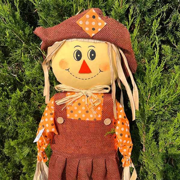 Close up of the face of a smiling scarecrow on a bamboo stake wearing an orange patchwork hat and dress with green legs.