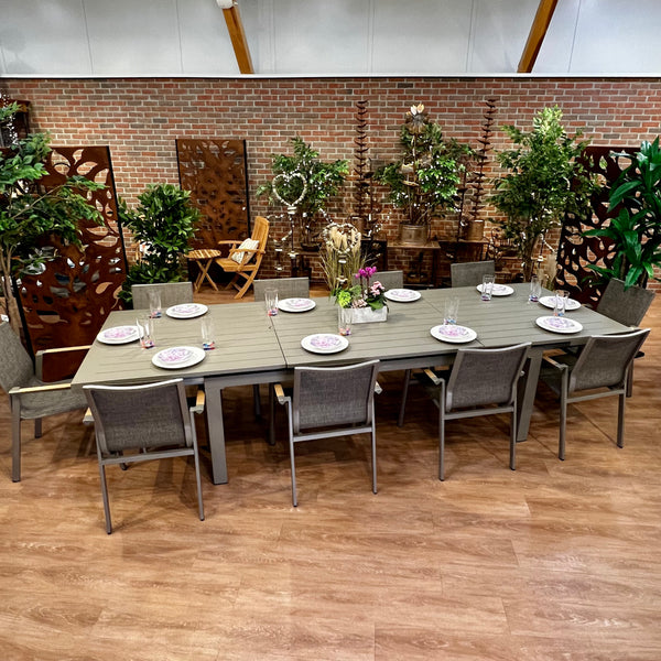 The Aspect Slat Outdoor Patio Extension Dining Set - 11 pc. by Delivery Only exudes modern elegance with its gray-finished table and eight chairs. A brick wall, wooden panels, and potted plants create a cozy indoor ambiance that evokes the feel of dining outdoors.