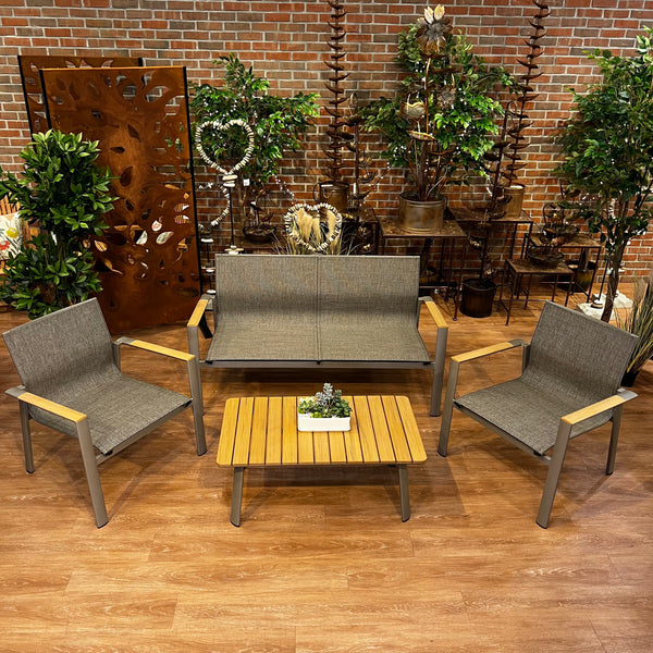 The Delivery Only Aspect Patio 4-piece Set includes a loveseat, coffee table, and chairs, featuring durable materials with gray cushions and wooden armrests. Its modern elegance is enhanced by a brick wall and decorative green plants.
