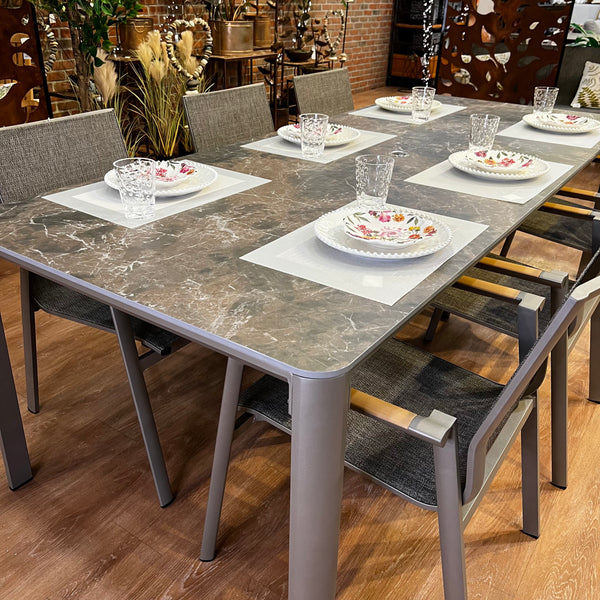 The Aspect Outdoor Patio Dining Set by Delivery Only, a 7-piece set, features a marble-like top and sleek gray chairs epitomizing modern elegance. Each setting includes floral-designed white plates beside clear glasses, set against brick walls and decorative plants for the ultimate outdoor dining bliss.