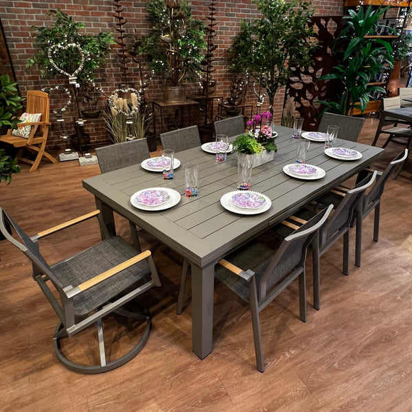 The Aspect Slat Outdoor Patio Extension Dining Set - 9 pc. by Delivery Only sits amid lush greenery. The set includes a large gray table with eight chairs, white floral plates, clear glasses, and a centerpiece of bright purple flowers on the wooden patio floor.