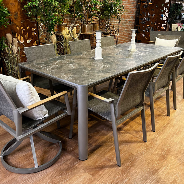Enhance your dining space with the Aspect Outdoor Patio Dining Set - 9 pc. from Delivery Only. A long marble table and six cushioned chairs create a chic, comfortable atmosphere, while two candlesticks and decorative plants add charm against a brick wall backdrop.