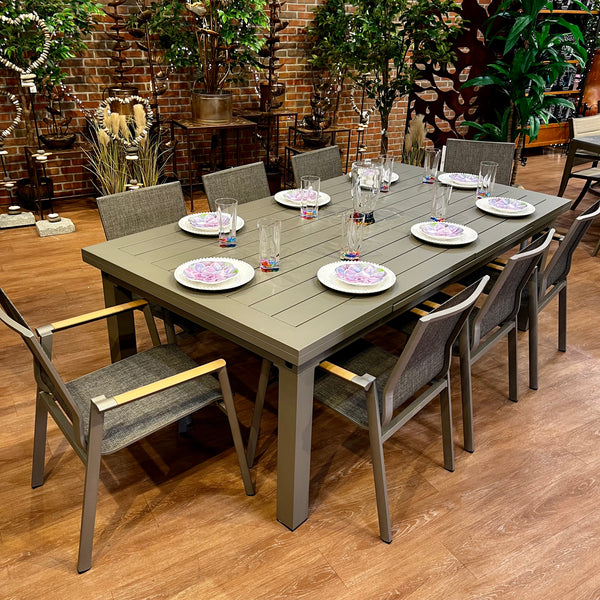 The Aspect Slat Outdoor Patio Extension Dining Set - 9 pc. by Delivery Only is arranged on a wooden floor and features eight elegant chairs. Each place is set with white floral plates, glasses, and cutlery. Leafy plants in pots against a brick wall create a serene backdrop.
